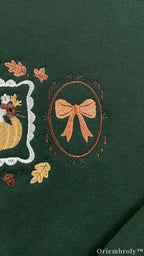 Close-up of an embroidered orange bow tied inside an oval frame on a dark green crewneck sweatshirt.