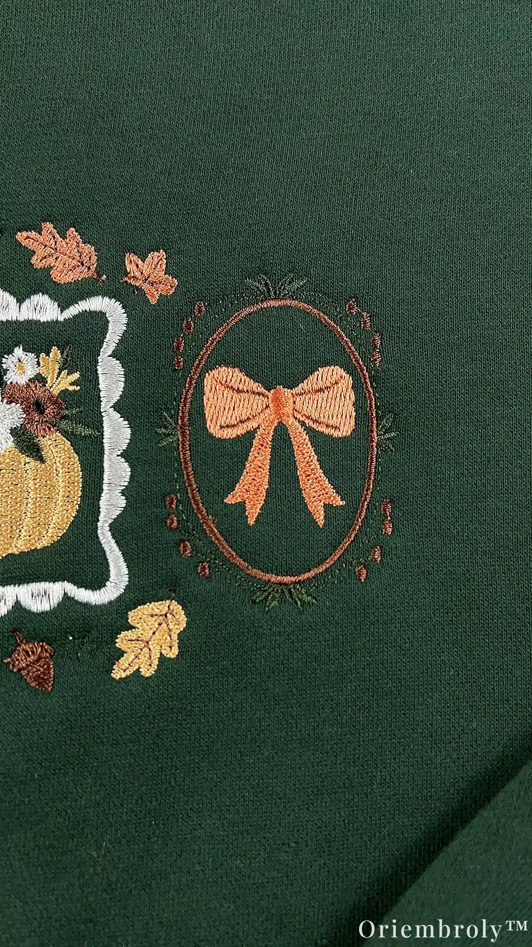 Close-up of an embroidered orange bow tied inside an oval frame on a dark green crewneck sweatshirt.
