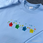Cozy baby blue crewneck with minimalist rainbow Junimo embroidery, perfect for Stardew Valley fans