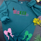 Teal sweatshirt with 'WICKED' text and decorative elements on a dark surface with flowers and leaves.