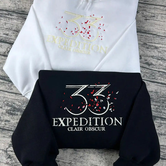 White and black hoodies with '33 Expedition Clair Obscur' logo on a wooden surface