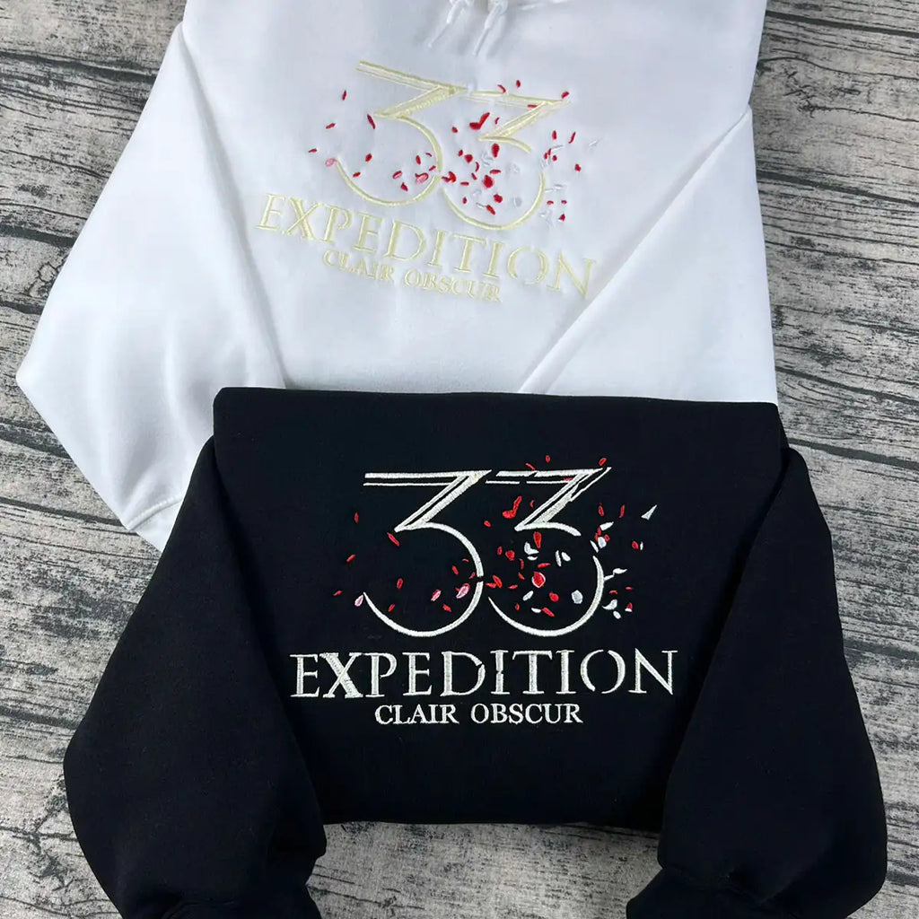 White and black hoodies with '33 Expedition Clair Obscur' logo on a wooden surface