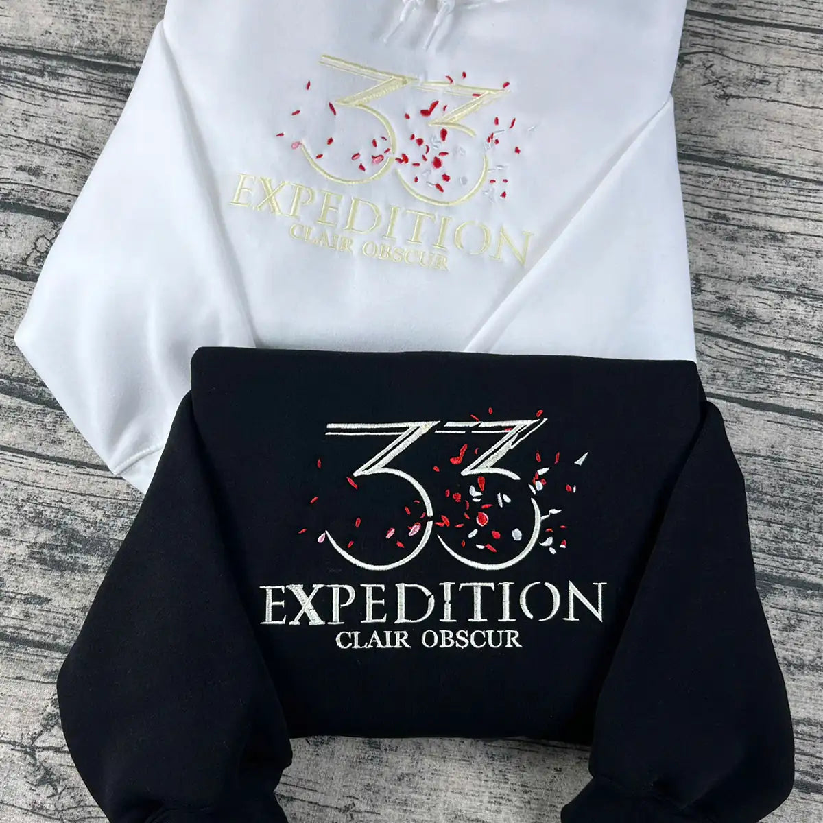 White and black hoodies with '33 Expedition Clair Obscur' logo on a wooden surface