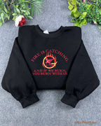 Flat lay of a black crewneck sweatshirt embroidered with red Fire Is Catching quote and Hunger Games Mockingjay pin logo