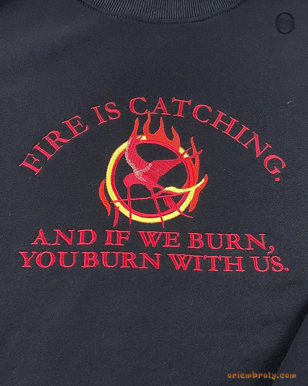 Close-up of red embroidered quote Fire Is Catching And If We Burn You Burn With Us with flaming Mockingjay symbol on a black sweater
