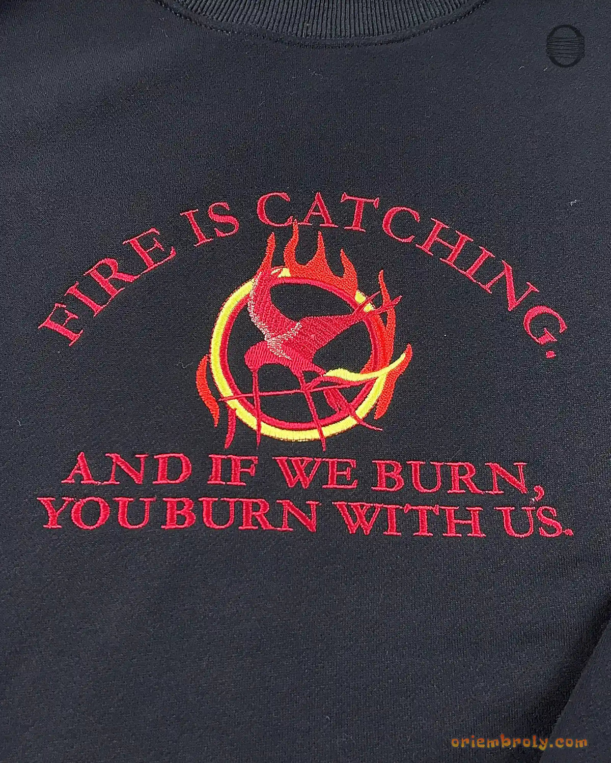 Close-up of red embroidered quote Fire Is Catching And If We Burn You Burn With Us with flaming Mockingjay symbol on a black sweater