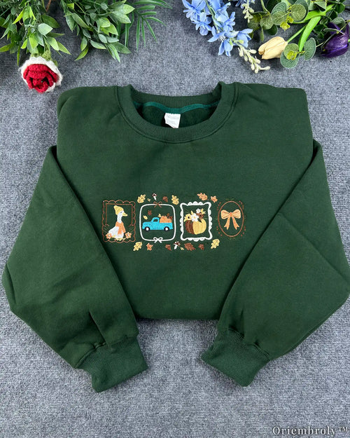 Flatlay view of a dark forest green crewneck sweatshirt embroidered with framed autumn icons including a duck wearing a scarf, a blue pickup truck with pumpkins, a fall pumpkin, and a bow.