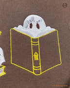 Close-up of a cute ghost hiding behind a giant open book embroidery detail on a brown sweatshirt