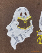 Close-up of a cute ghost holding and reading a open book embroidery on a brown sweater