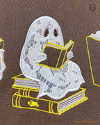 Close-up of a ghost sitting on a stack of books reading embroidery detail on a brown sweatshirt