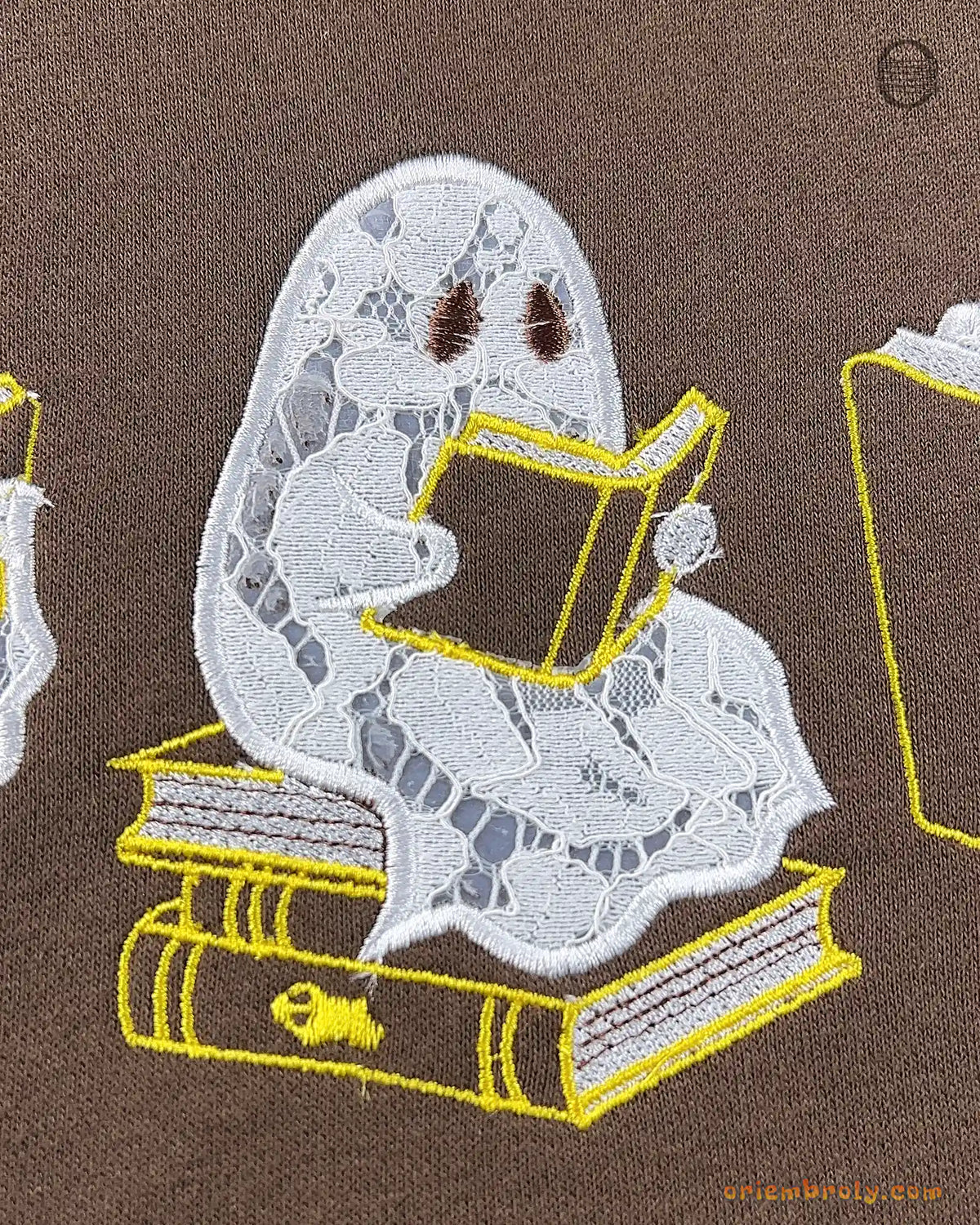 Close-up of a ghost sitting on a stack of books reading embroidery detail on a brown sweatshirt