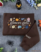 Flat lay of a brown Gilmore Girls embroidered sweatshirt with Luke's diner coffee cup, pumpkin, and autumn details