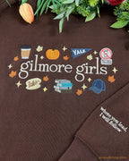 Close-up of Gilmore Girls autumn embroidery featuring a coffee cup, pumpkin, Yale pennant, and books on a brown sweater