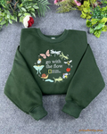 Flat lay of a forest green crewneck sweatshirt embroidered with Go With The Flow and nature insect wreath