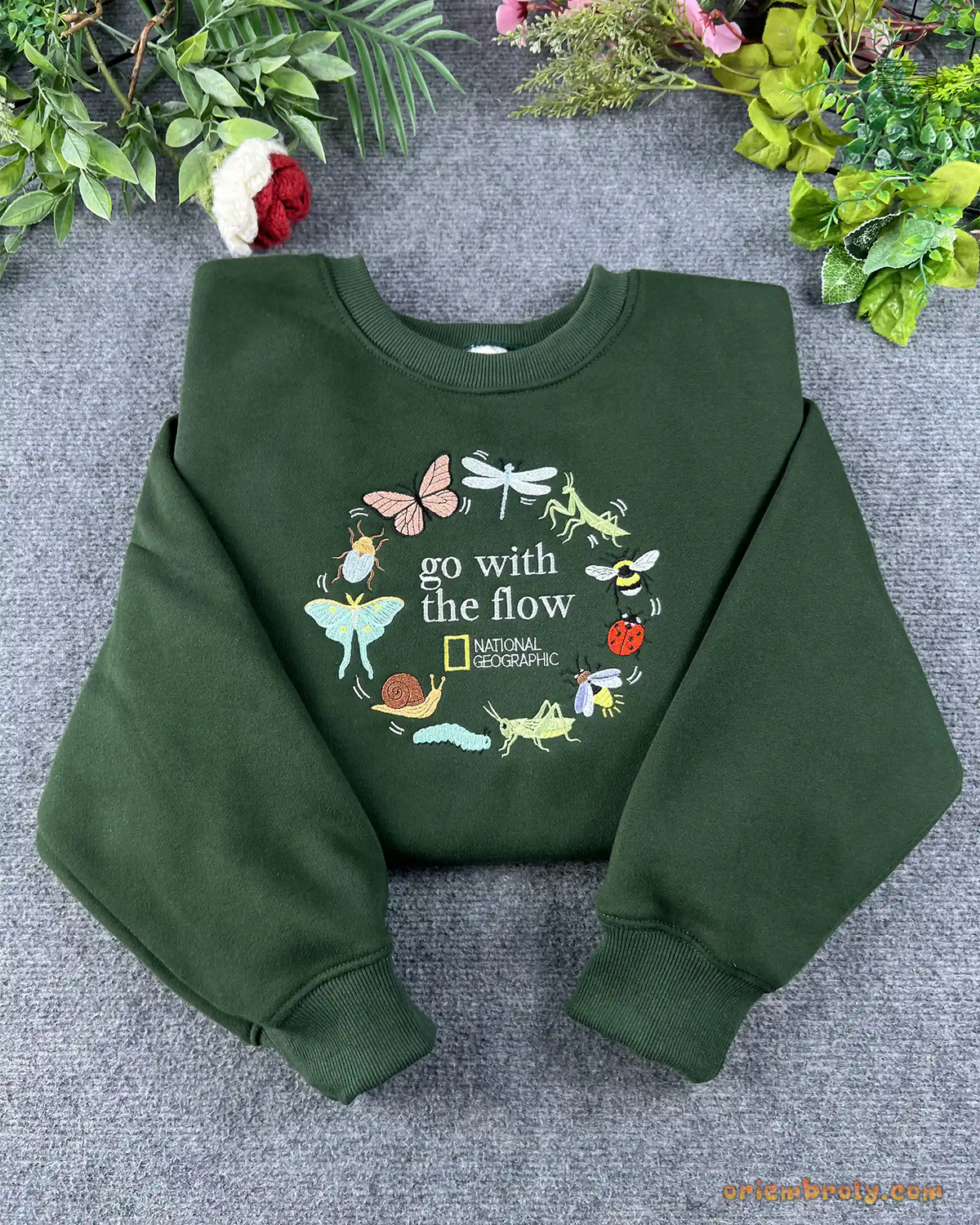 Flat lay of a forest green crewneck sweatshirt embroidered with Go With The Flow and nature insect wreath