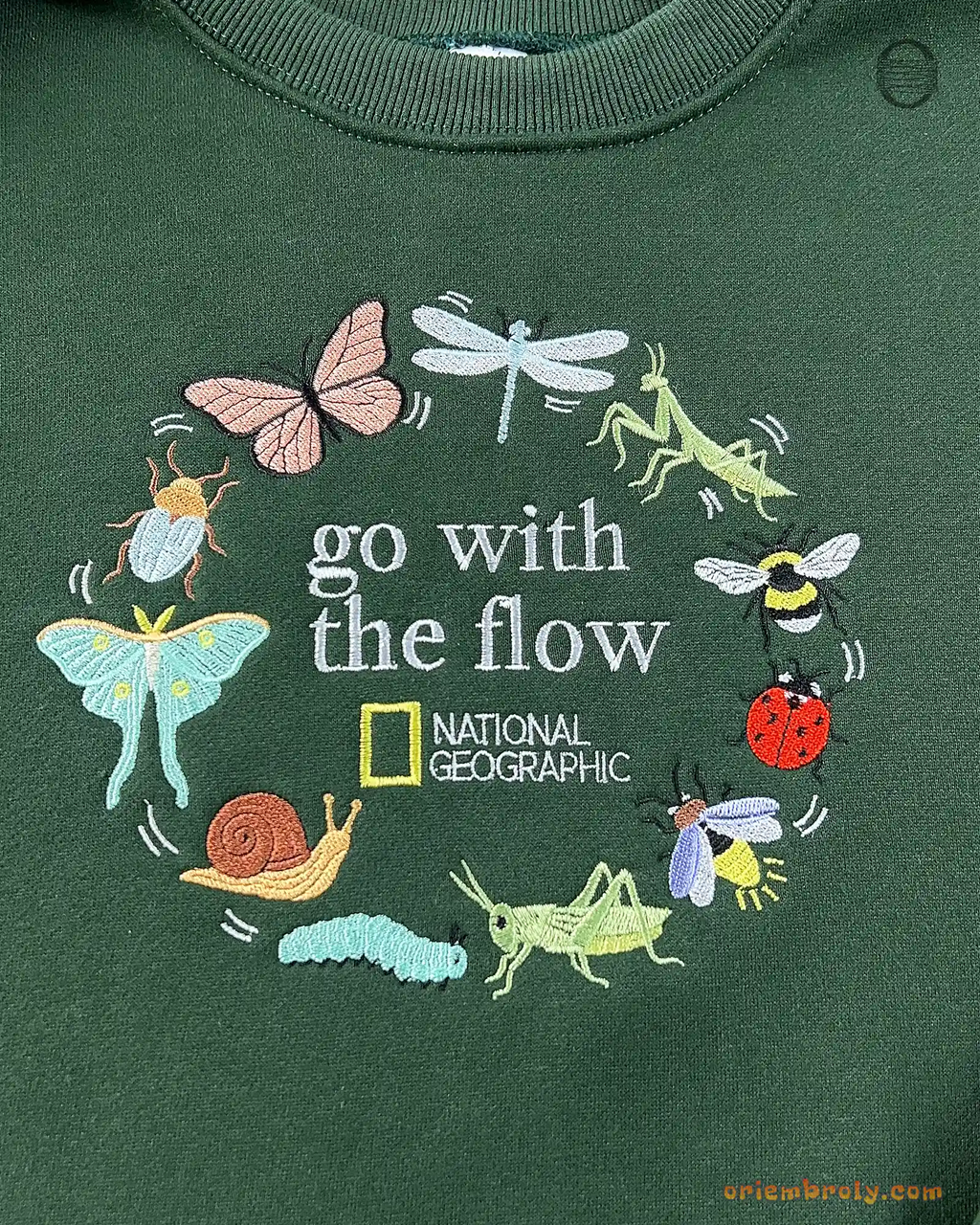 Close-up of Go With The Flow embroidery featuring butterflies, dragonflies, snails, and beetles wreath on green sweater