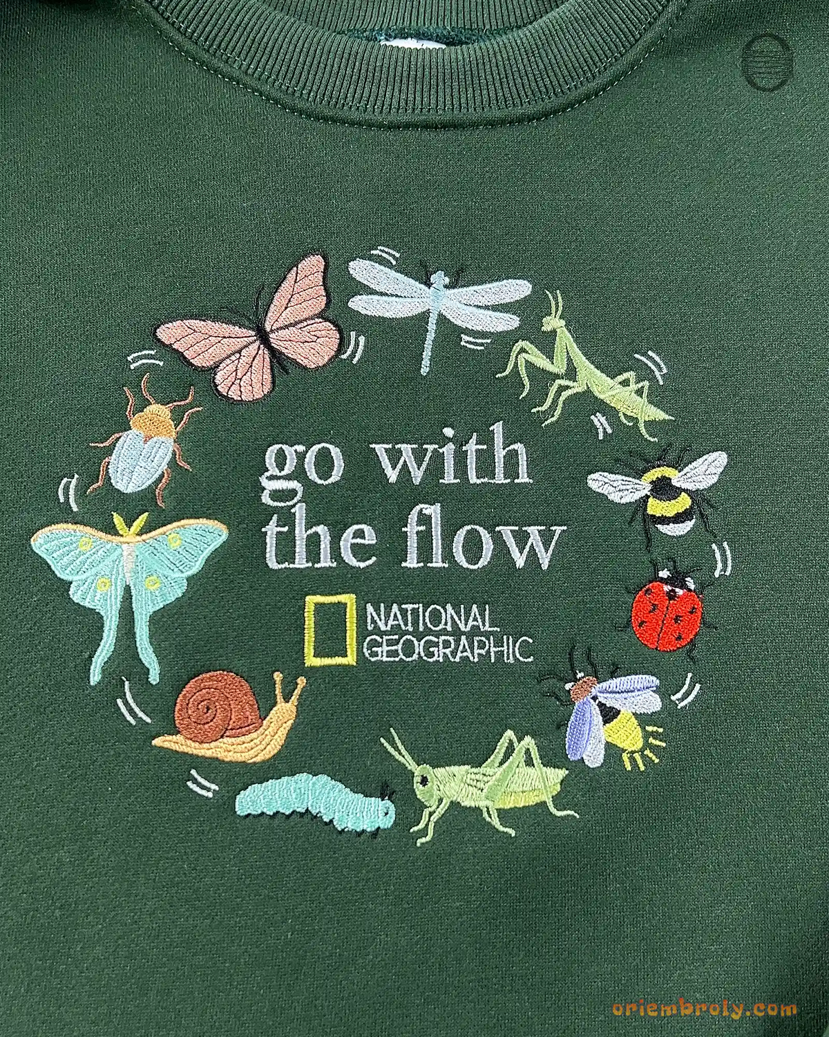 Close-up of Go With The Flow embroidery featuring butterflies, dragonflies, snails, and beetles wreath on green sweater