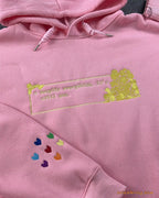 Close-up of gold embroidered quote with hearts box and colorful rainbow hearts sleeve detail on a pink hoodie