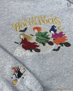 Close-up of ash gray sweatshirt embroidered with A Little Hocus Pocus text, colorful witch hair, and flying broomsticks.
