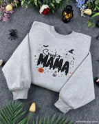 Angled view of a gray Spooky MAMA embroidered Halloween sweatshirt with festive autumn decorations around it.