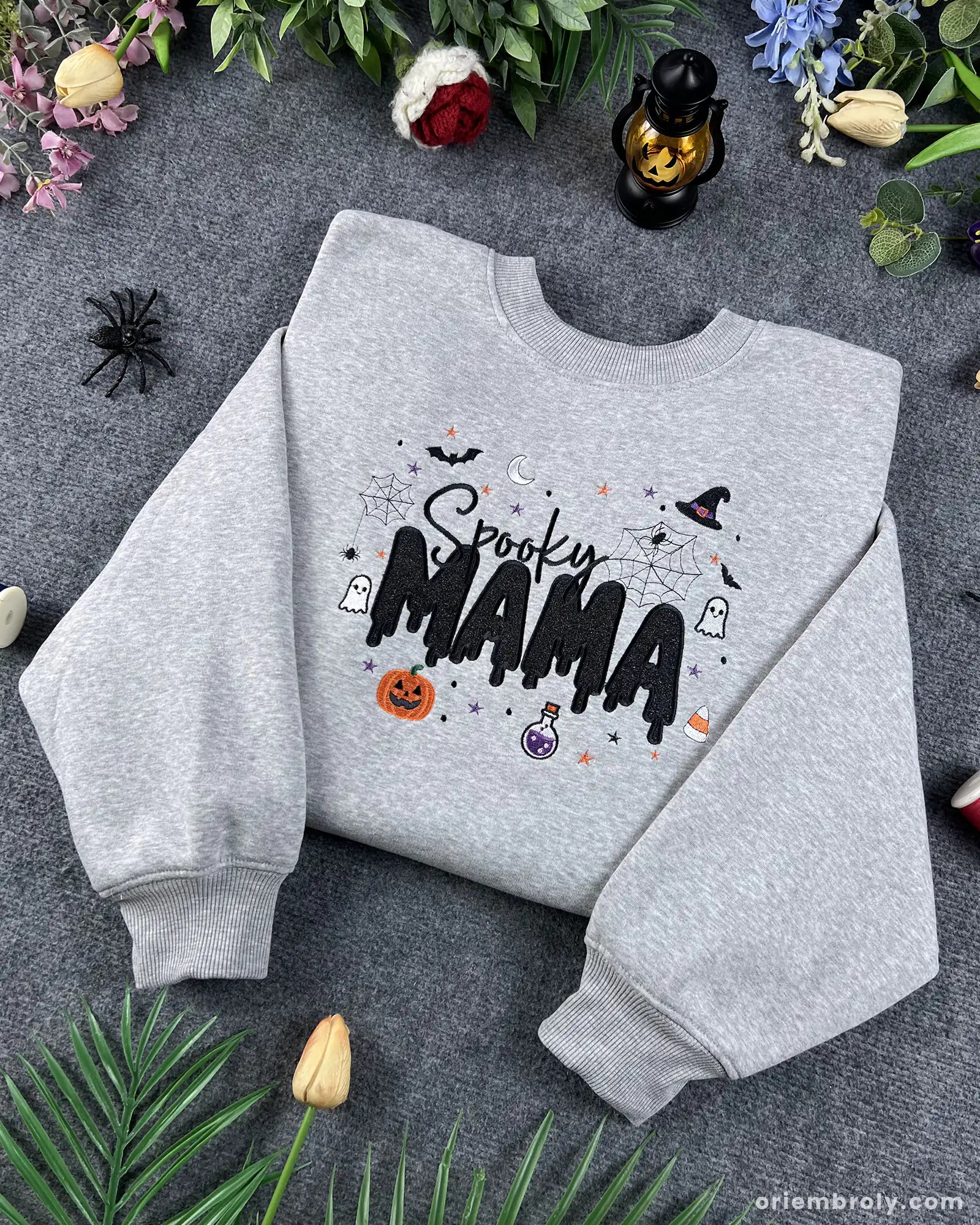 Angled view of a gray Spooky MAMA embroidered Halloween sweatshirt with festive autumn decorations around it.