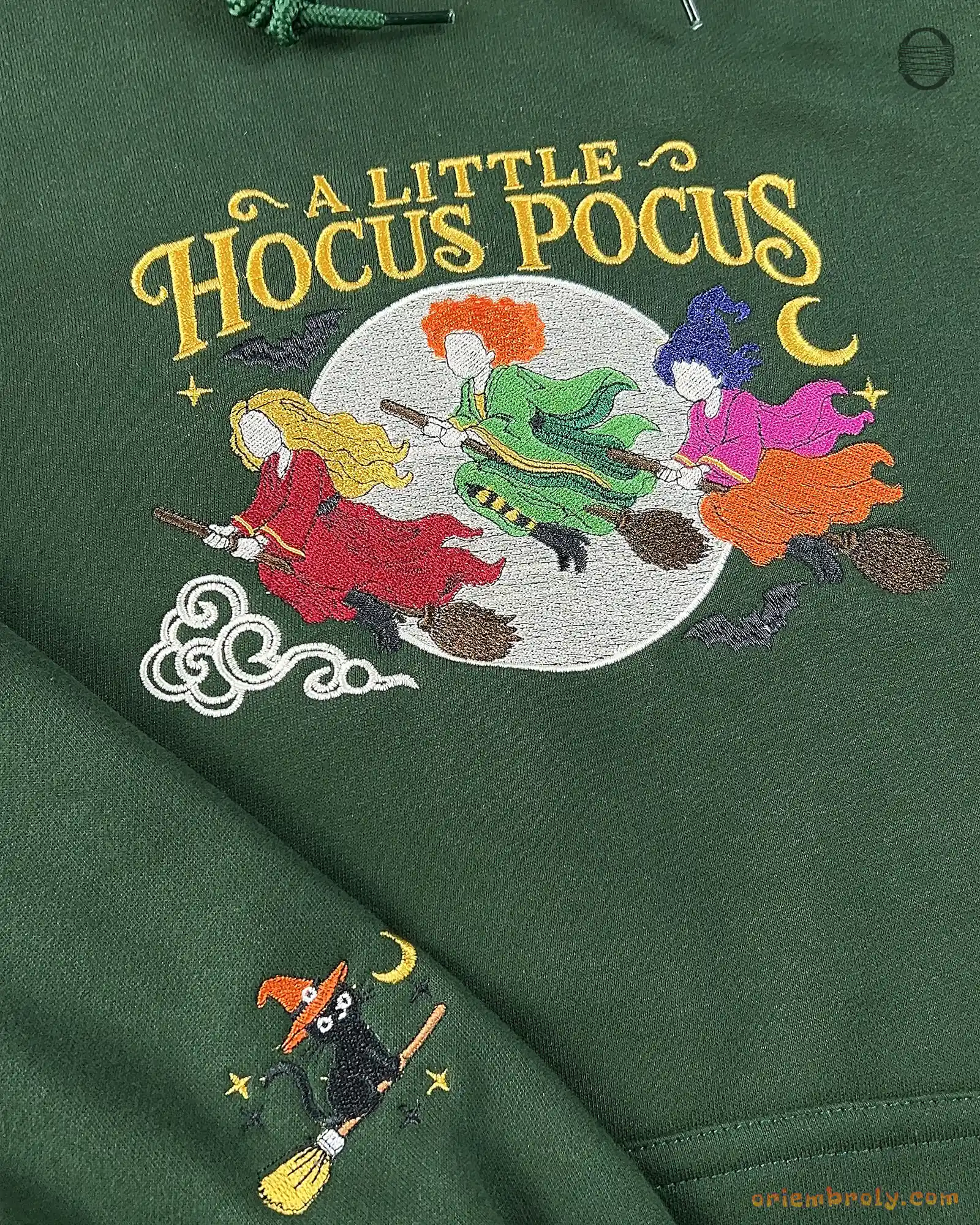 Close-up of green hoodie featuring A Little Hocus Pocus embroidery with three witches riding broomsticks and pumpkin sleeve print.
