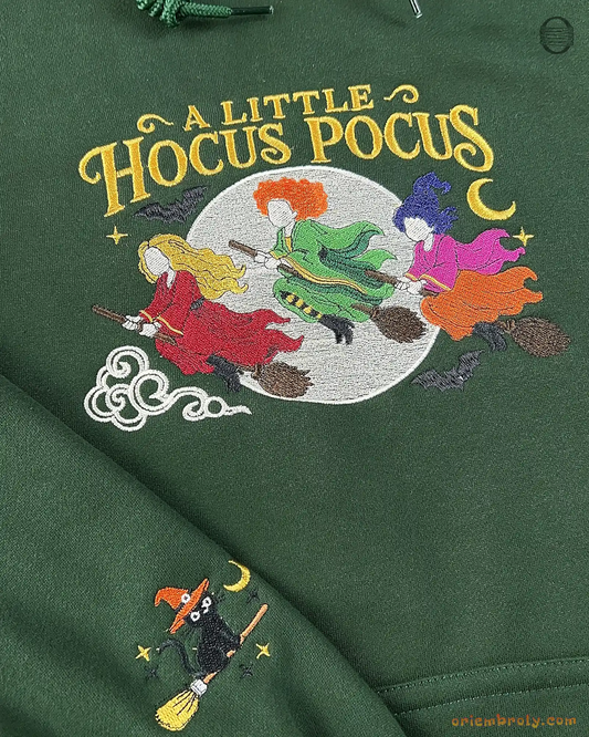 Close-up of green hoodie featuring A Little Hocus Pocus embroidery with three witches riding broomsticks and pumpkin sleeve print.
