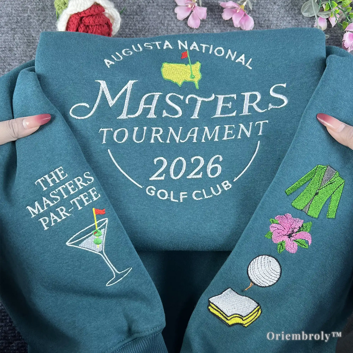 Close-up of embroidered icons including the iconic green jacket, pink azalea, and golf ball on a teal Masters 2026 pullover. Detailed high-density satin stitching