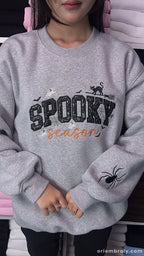 Front view of model wearing heather grey spooky season embroidered sweatshirt with black sequin letters.