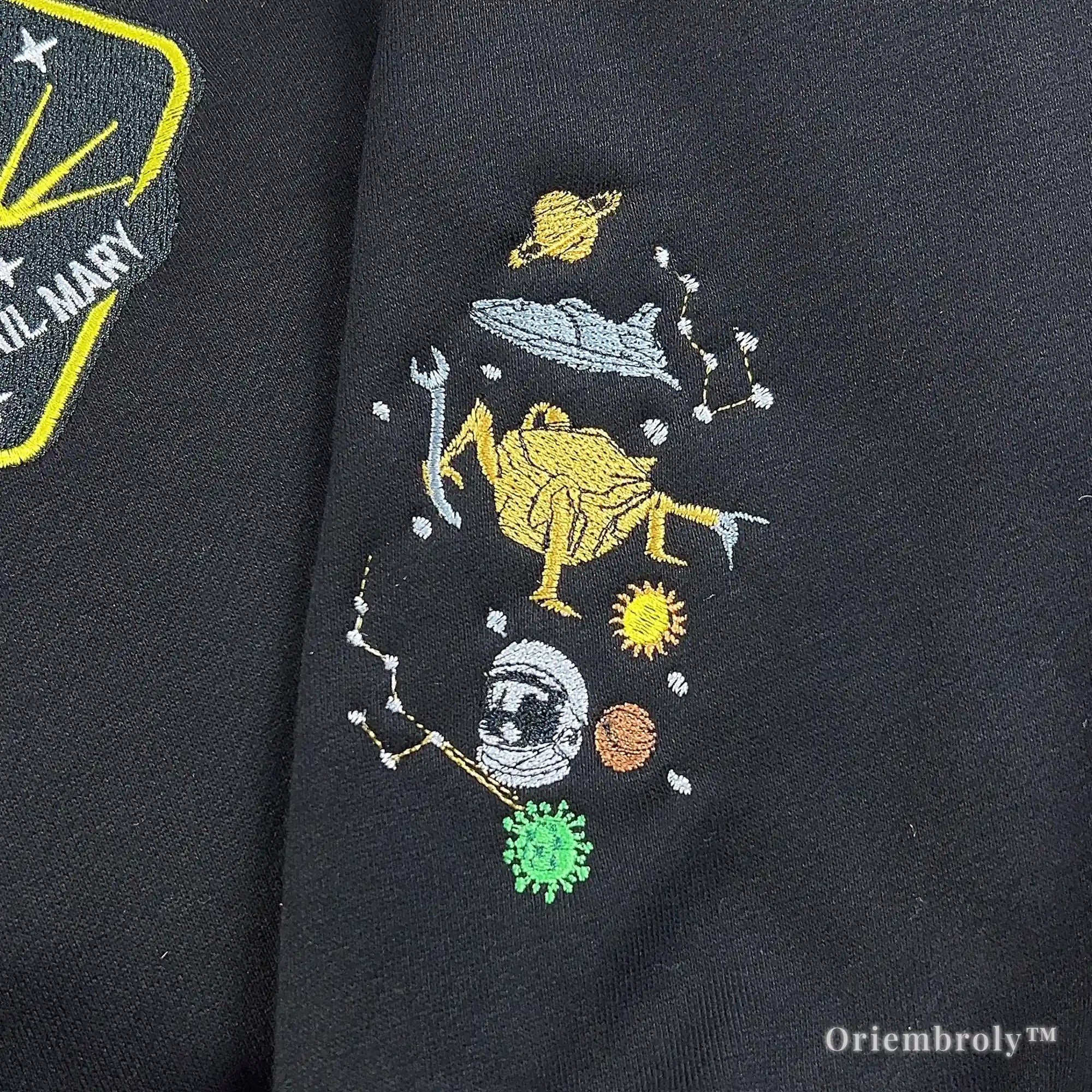 Close-up of custom high-density satin stitch embroidery on a black heavyweight quarter-zip. Features the Hail Mary mission logo and cosmic constellations in vibrant yellow and white thread.