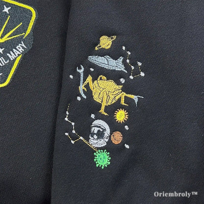 Close-up of custom high-density satin stitch embroidery on a black heavyweight quarter-zip. Features the Hail Mary mission logo and cosmic constellations in vibrant yellow and white thread.