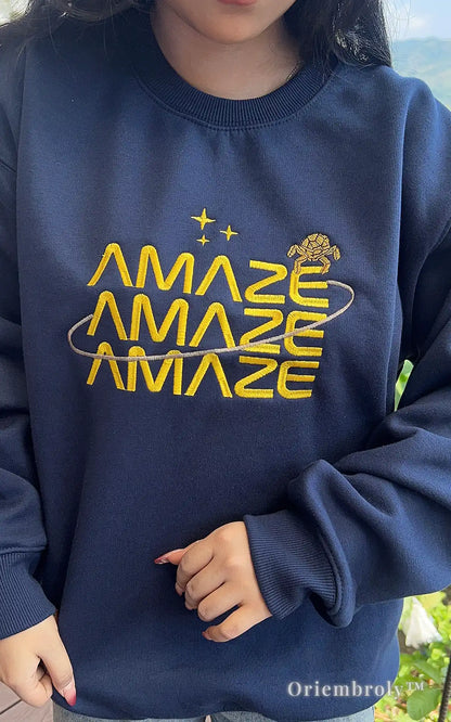 Close-up of a heavyweight navy crewneck with vibrant yellow "Amaze Amaze Amaze" embroidery. Durable, high-quality thread work on soft fleece for a premium streetwear experience