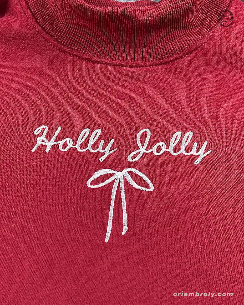Close-up of white Holly Jolly cursive script and ribbon bow embroidered on the chest of a maroon red sweatshirt