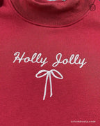 Close-up of white Holly Jolly cursive script and ribbon bow embroidered on the chest of a maroon red sweatshirt