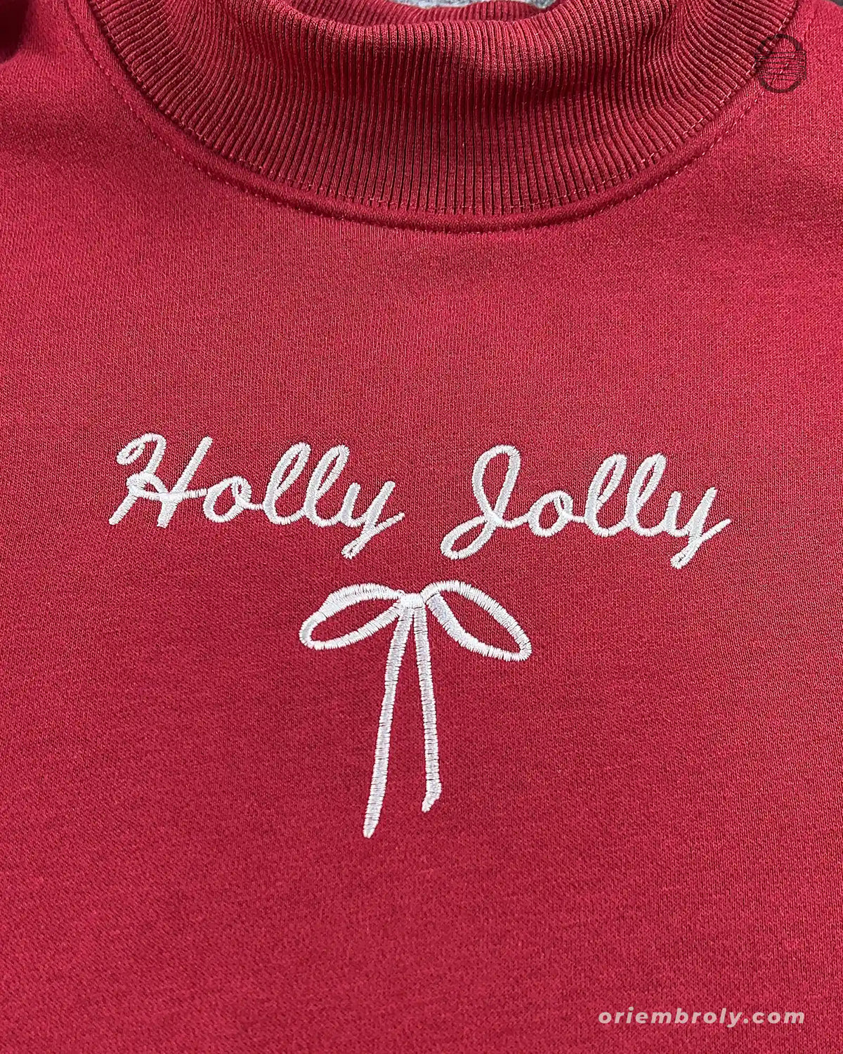 Close-up of white Holly Jolly cursive script and ribbon bow embroidered on the chest of a maroon red sweatshirt