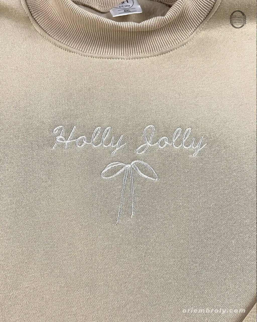 Close-up of subtle white Holly Jolly embroidery with a delicate bow on a sand beige crewneck sweater