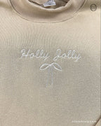 Close-up of subtle white Holly Jolly embroidery with a delicate bow on a sand beige crewneck sweater