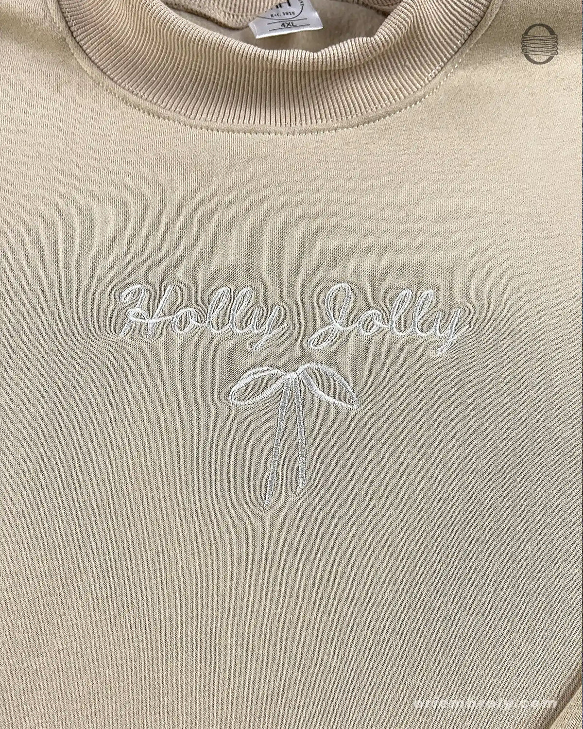 Close-up of subtle white Holly Jolly embroidery with a delicate bow on a sand beige crewneck sweater