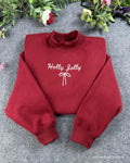 Flat lay of a maroon red crewneck sweatshirt embroidered with white Holly Jolly cursive text and a delicate coquette ribbon bow