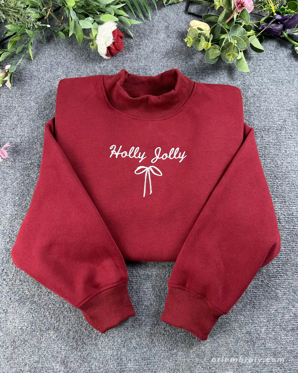 Flat lay of a maroon red crewneck sweatshirt embroidered with white Holly Jolly cursive text and a delicate coquette ribbon bow