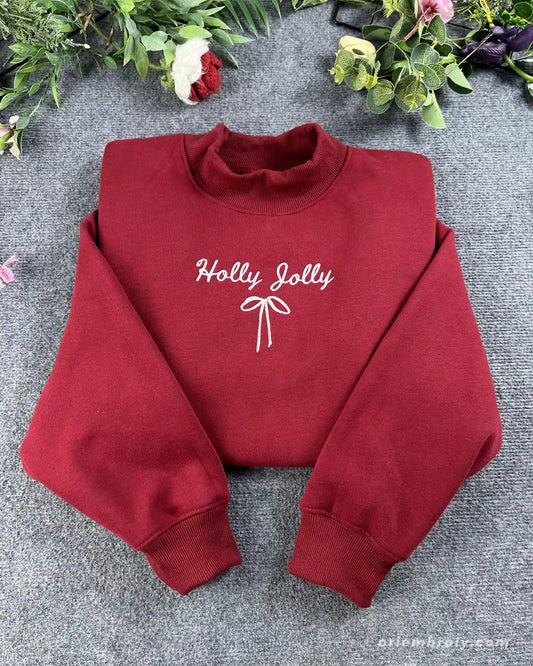 Flat lay of a maroon red crewneck sweatshirt embroidered with white Holly Jolly cursive text and a delicate coquette ribbon bow