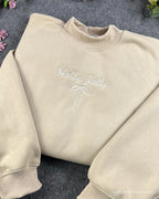 Flat lay of a sand beige crewneck sweatshirt embroidered with white Holly Jolly text and minimal coquette bow detail