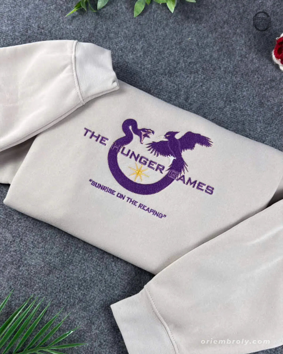 Close-up of purple and gold embroidered Mockingjay symbol and Sunrise on the Reaping title on a sand sweatshirt