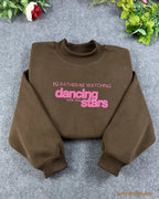 Flat lay of a chocolate brown crewneck sweatshirt embroidered with hot pink I'd Rather Be Watching Dancing with the Stars text