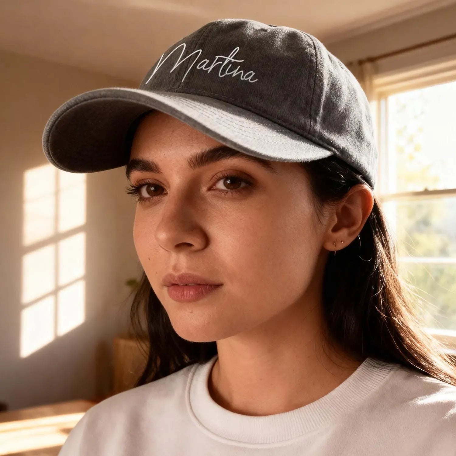 Woman wearing a cap with 'Martina' embroidered on it, sitting indoors by a window.