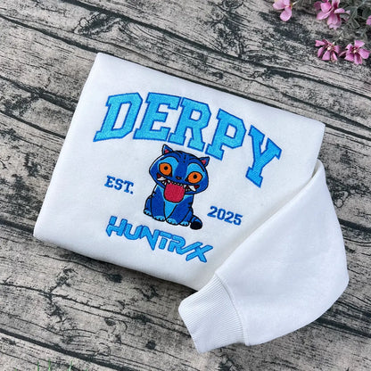White sweatshirt with 'DERPY' and cartoon character on a wooden surface with flowers.