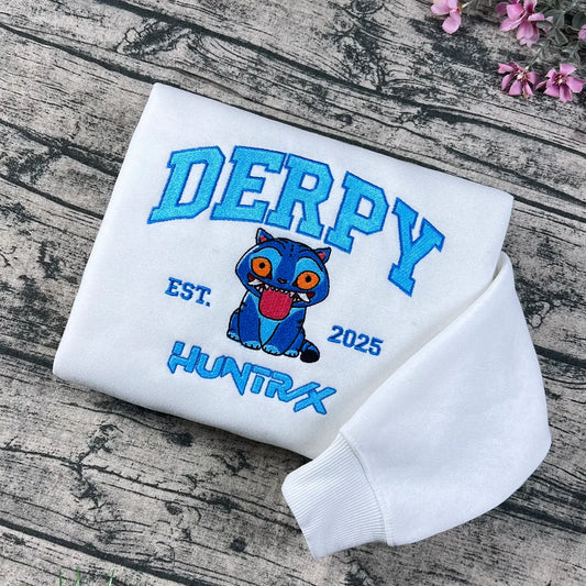 White sweatshirt with 'DERPY' and cartoon character on a wooden surface with flowers.