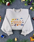 Flatlay view of an oatmeal beige crewneck sweatshirt with embroidered La La La Season text, autumn leaves, coffee cup, and cozy fall icons.