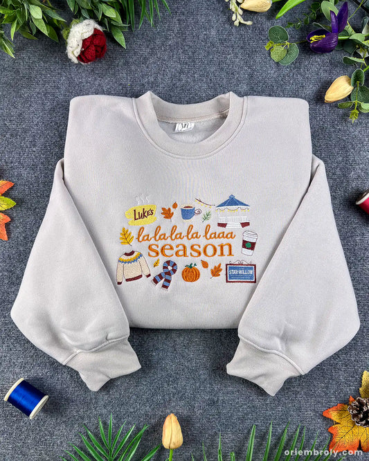 Flatlay view of an oatmeal beige crewneck sweatshirt with embroidered La La La Season text, autumn leaves, coffee cup, and cozy fall icons.