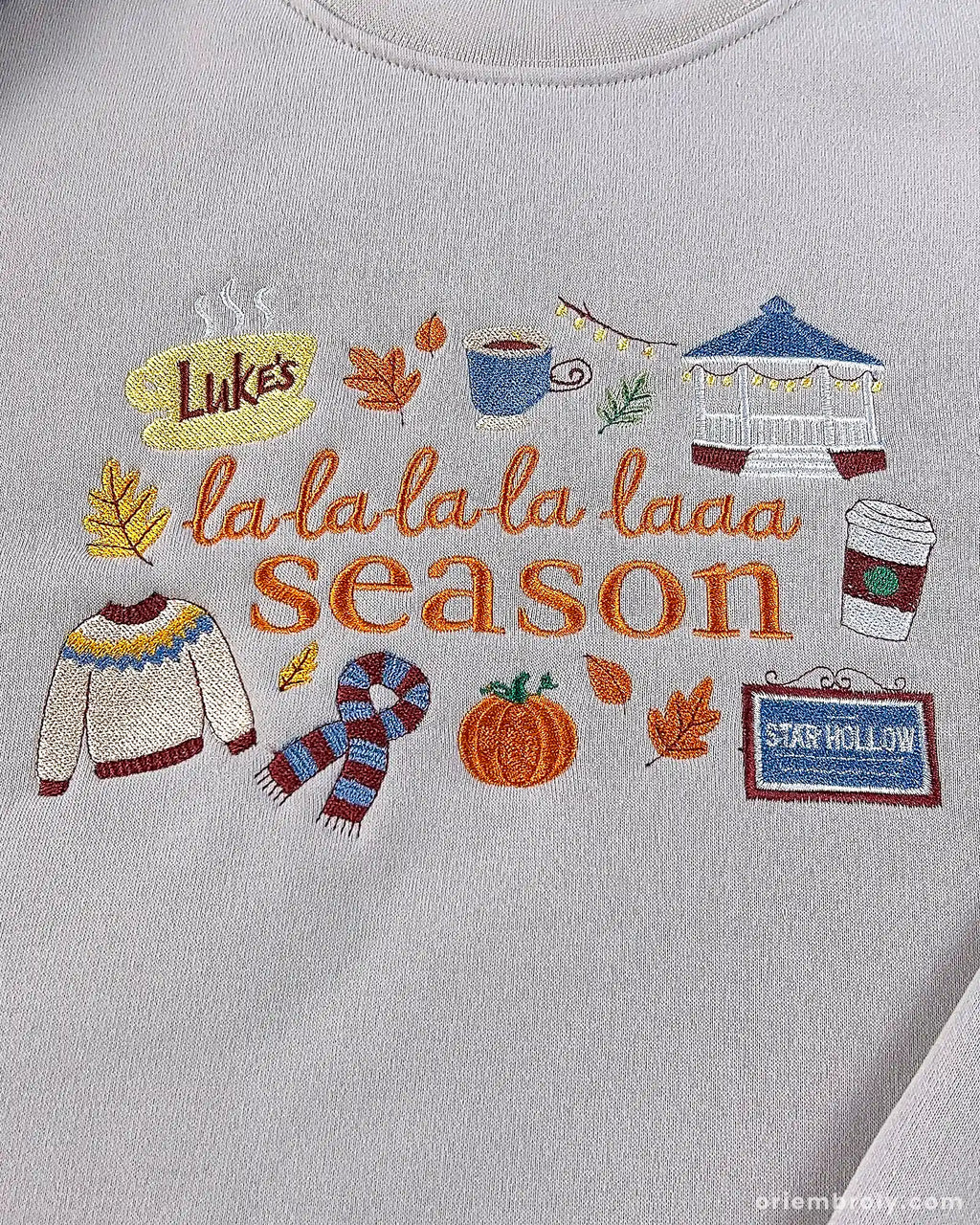 Close-up of fine embroidery on a beige sweatshirt showing La La La Season lettering surrounded by pumpkin, scarf, and warm coffee mug.